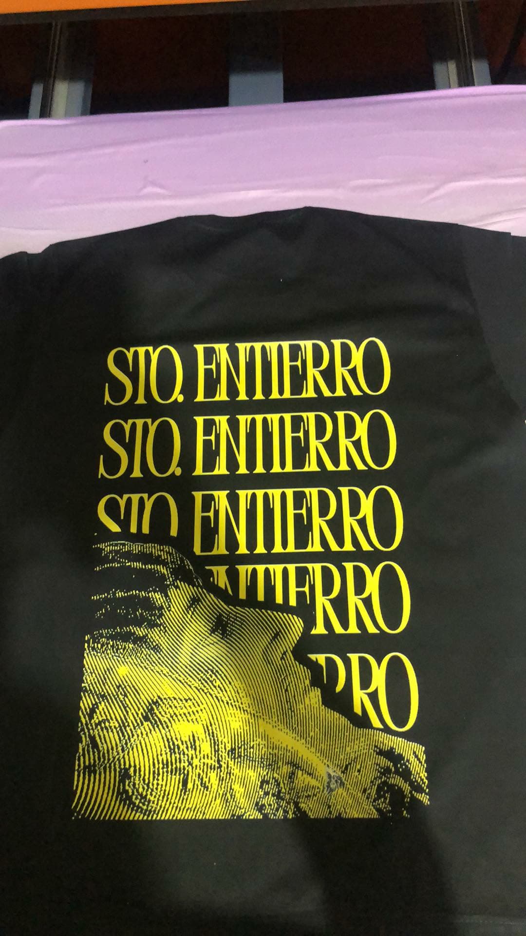 Back of a black tee printed with five stacked yellow "Sto. Entierro" lines over a halftone yellow image